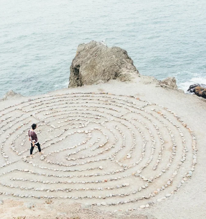A woman walking around a huge mandala drawn in the sands of a beach.