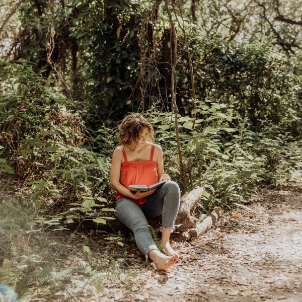 Alison sitting on the ground, reading a book in a sunny day in the woods.